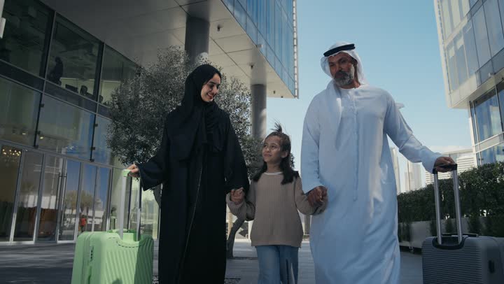 الاستعداد للسفر في جولة سياحية، قضاء رحلة صيفية ممتعة، مفهوم السياحة في إجازة الصيف، رجل عربي خليجي اماراتي يرتدي الكندورة والغترة يقف برفقة زوجته وابنته امام الفندق، تجهيز حقائب السفر والامتعة، ايماءات السعادة والسرور