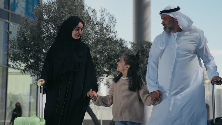 Preparing to travel on a tourist trip, spending a pleasant summer trip, the concept of tourism during the summer vacation, an Arab Gulf Emirati man wearing a kandura and ghutra standing with his wife and daughter in front of the hotel, preparing travel bags and luggage, gestures of happiness and pleasure