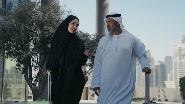 قضاء رحلة صيفية مسلية، مفهوم السياحة في إجازة الصيف، زوجان عربيان خليجيان اماراتيان يجران حقائب الامتعة امام الفندق، تعابير وايماءات السعادة والسرور، عائلة تقضي وقت ممتع، الاستعداد للسفر في جولة سياحية