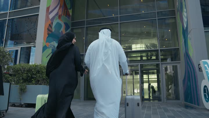 مفهوم السياحة في إجازة الصيف، تصوير من الخلف لزوجين عربيان خليجيين اماراتيين يجران حقائب الامتعة امام الفندق، عائلة سعودية تقضي وقت ممتع، الاستعداد للسفر في جولة سياحية، قضاء رحلة صيفية ممتعة ومسلية