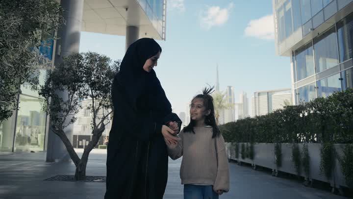تبادل الأحاديث والأخبار بإيماءات السرور والفرح, عائلة سعودية تقوم بالتنزه في وقت العطلة في إحدى الاماكن العامة في الإمارات, أم عربية خليجية إماراتية ترتدي الحجاب والعباءة تقوم بالمشي والتجول في الهواء الطلق برفقة ابنتها، الاشارة إلى شيء ما، تقديم الاهتمام للأبناء