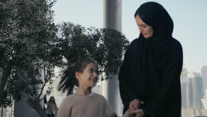 تقديم الاهتمام والرعاية للأبناء، تبادل الأحاديث والأخبار بإيماءات السرور والفرح, عائلة سعودية تقوم بالتنزه في وقت العطلة في إحدى الاماكن العامة في الإمارات, أم عربية خليجية إماراتية ترتدي الحجاب والعباءة تقوم بالمشي والتجول في الهواء الطلق برفقة ابنتها