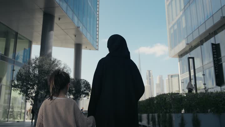 تقديم الاهتمام والرعاية للأبناء، تبادل الأحاديث بإيماءات السرور والفرح, عائلة سعودية تقوم بالتنزه في وقت العطلة في إحدى الاماكن العامة في الإمارات, تصوير من الخلف لأم عربية خليجية إماراتية ترتدي الحجاب والعباءة تقوم بالمشي والتجول في الهواء الطلق برفقة ابنتها