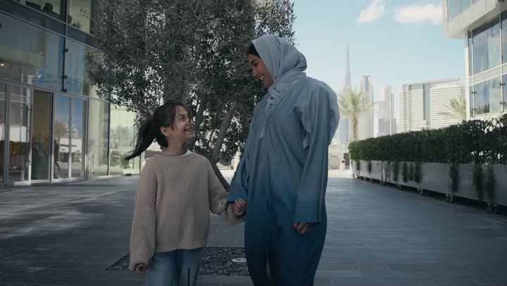 A Saudi family is out for a walk during the holiday in a public place in the Emirates, exchanging conversations and news with gestures of pleasure and joy. An Emirati Gulf Arab mother wearing a hijab and abaya is walking and strolling in the open air with her daughter, pointing to something, giving attention to the children.