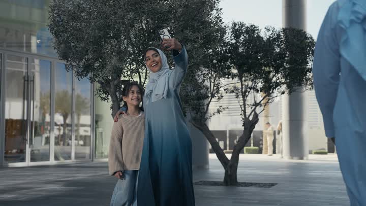 أم عربية خليجية إماراتية ترتدي الحجاب والعباءة تقوم بالتجول في الهواء الطلق برفقة ابنتها، التنزه في وقت العطلة في إحدى الاماكن العامة في الإمارات, امرأة سعودية تقوم بالتقاط صور السيلفي التذكارية باستخدام الهاتف المحمول بإيماءات السعادة والسرور