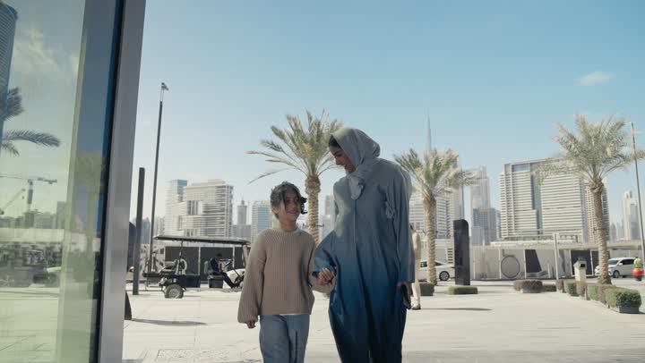 عائلة سعودية تقوم بالتنزه في وقت العطلة في إحدى الاماكن العامة في الإمارات, تبادل الأحاديث والأخبار بإيماءات السرور والفرح, أم عربية خليجية إماراتية ترتدي الحجاب والعباءة تقوم بالمشي والتجول في الهواء الطلق برفقة ابنتها، الاشارة إلى شيء ما، تقديم الاهتمام للأبناء