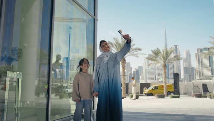 An Emirati Gulf Arab mother wearing a hijab and abaya walks outdoors with her daughter, taking a walk during the holiday in a public place in the Emirates. A Saudi woman takes souvenir selfies using her mobile phone with gestures of happiness and pleasure.