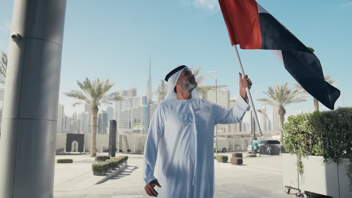 رفع راية البلاد للاعلى، الاحتفال بذكرى تأسيس الاتحاد 2 ديسمبر في الطريق، التجول في الهواء الطلق، يوم العلم 3 نوفمبر، حب الوطن والاعتزاز به، اليوم الوطني الاماراتي 1971، رجل عربي خليجي اماراتي يرتدي الكندورة والغترة يمسك علم الامارات العربية المتحدة في النهار