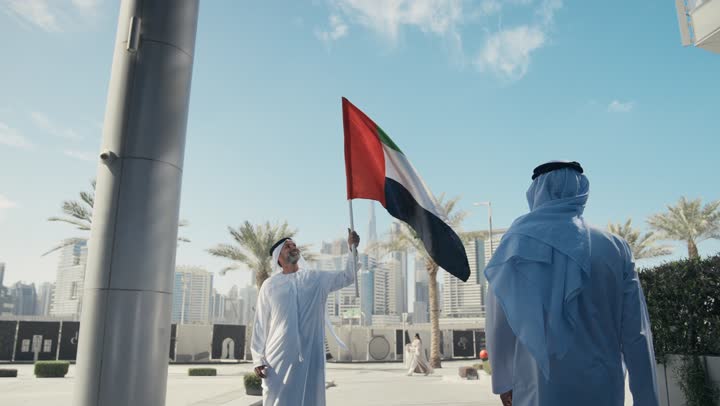 Raising the country's flag high, celebrating the anniversary of the founding of the Union on December 2nd on the road, walking in the open air, Flag Day on November 3rd, love and pride of the homeland, UAE National Day 1971, an Arab Gulf Emirati man wearing a kandura and ghutra holding the flag of the United Arab Emirates during the day