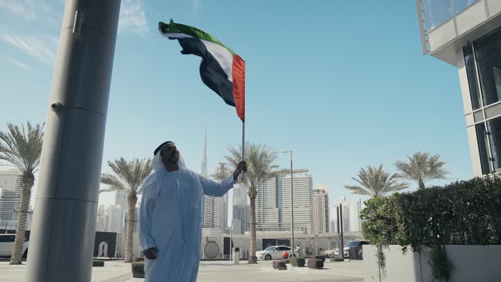 رفع راية البلاد للاعلى، الاحتفال بذكرى تأسيس الاتحاد 2 ديسمبر في الطريق، التجول في الهواء الطلق، يوم العلم 3 نوفمبر، حب الوطن والاعتزاز به، اليوم الوطني الاماراتي 1971، رجل عربي خليجي اماراتي يرتدي الكندورة والغترة يمسك علم الامارات العربية المتحدة في النهار