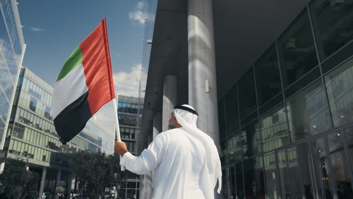 رفع راية البلاد للاعلى، الاحتفال بذكرى تأسيس الاتحاد 2 ديسمبر في الطريق، التجول في الهواء الطلق، يوم العلم 3 نوفمبر، حب الوطن والاعتزاز به، اليوم الوطني الاماراتي 1971، تصوير من الخلف لرجل عربي خليجي اماراتي يرتدي الكندورة والغترة يمسك علم الامارات العربية المتحدة