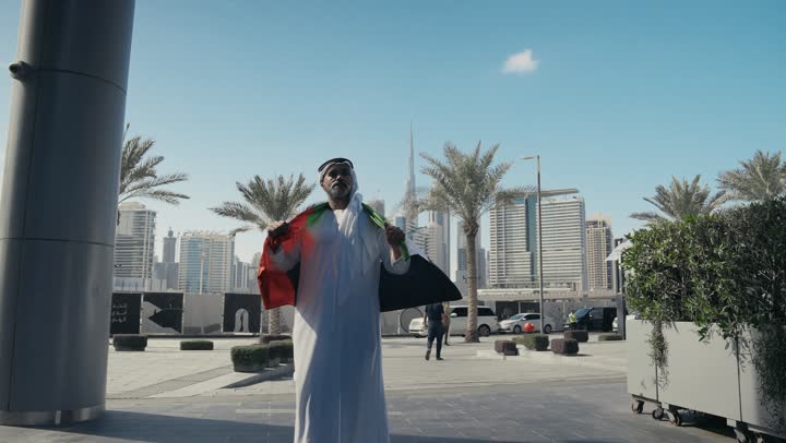 الاحتفال بذكرى تأسيس الاتحاد 2 ديسمبر في الطريق، التجول في الهواء الطلق، يوم العلم 3 نوفمبر، حب الوطن والاعتزاز به، اليوم الوطني الاماراتي 1971، رجل عربي خليجي اماراتي يرتدي الكندورة والغترة التقليدية يضع علم الامارات العربية المتحدة على كتفيه في النهار
