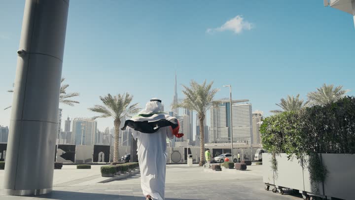 حب الوطن والاعتزاز به، الاحتفال بذكرى تأسيس الاتحاد 2 ديسمبر في الطريق، التجول في الهواء الطلق، يوم العلم 3 نوفمبر، اليوم الوطني الاماراتي 1971، تصوير من الخلف لرجل عربي خليجي اماراتي يرتدي الكندورة والغترة يضع علم الامارات العربية المتحدة على كتفيه في النهار