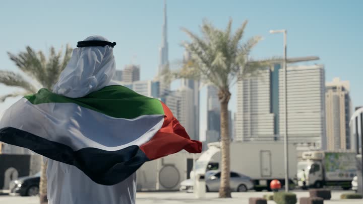 حب الوطن والاعتزاز به، الاحتفال بذكرى تأسيس الاتحاد 2 ديسمبر في الطريق، التجول في الهواء الطلق، يوم العلم 3 نوفمبر، اليوم الوطني الاماراتي 1971، تصوير من الخلف لرجل عربي خليجي اماراتي يرتدي الكندورة والغترة يضع علم الامارات العربية المتحدة على كتفيه في النهار