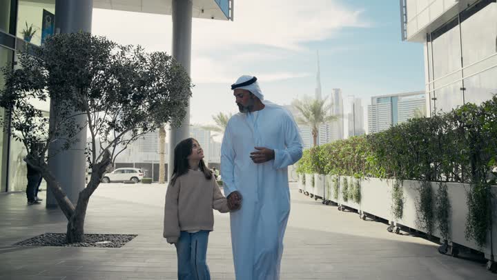 Exchanging conversations and news with gestures of pleasure and joy, an Emirati Gulf Arab father wearing a kandura and ghutra walking and strolling in the open air with his daughter, happy family memories and moments, a walk during the holiday in one of the public places in the Emirates