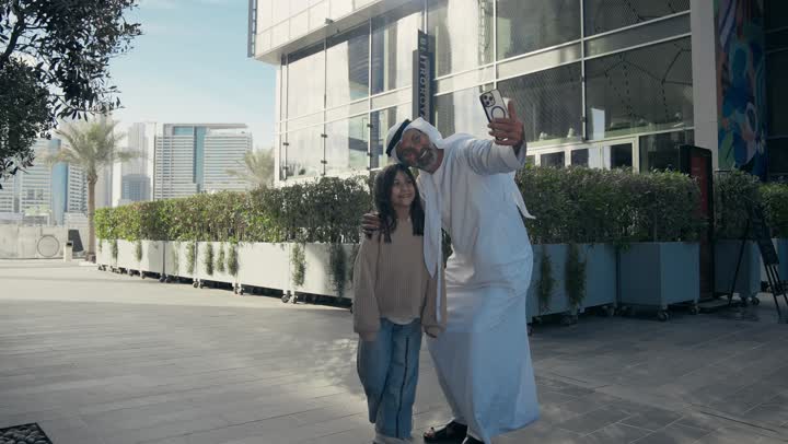 أب عربي خليجي إماراتي يرتدي الكندورة والغترة يقوم بالتجول في الهواء الطلق برفقة ابنته, الذكريات واللحظات العائلية السعيدة، التنزه في وقت العطلة في إحدى الاماكن العامة في الإمارات، رجل سعودي يقوم بالتقاط صور السيلفي التذكارية باستخدام الهاتف المحمول بإيماءات السعادة
