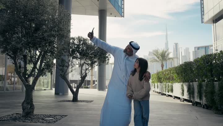 أب عربي خليجي إماراتي يرتدي الكندورة والغترة يقوم بالتجول في الهواء الطلق برفقة ابنته, الذكريات واللحظات العائلية السعيدة، التنزه في وقت العطلة في إحدى الاماكن العامة في الإمارات، رجل سعودي يقوم بالتقاط صور السيلفي التذكارية باستخدام الهاتف المحمول بإيماءات السعادة