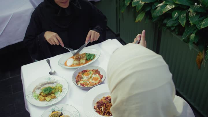 Sharing food with friends, two Emirati Gulf Arab women sitting at a table in a cafe, developing social relationships, chatting and having a good time with friends, an upscale coffee shop in the Emirates, expressions of happiness and pleasure at a gathering of friends, two Saudi friends exchanging conversation.