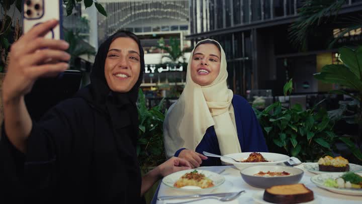 Taking souvenir photos, two Emirati Gulf Arab women sitting at a table in a cafe, developing social relations, a high-end coffee shop in the Emirates, expressions of happiness and pleasure at a gathering of friends, chatting and having a good time with friends, two Saudi friends exchanging talk.