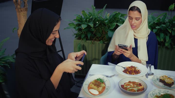 تطوير العلاقات الإجتماعية، كوفي شوب راقي بالامارات، تعابير السعادة والسرور بلمة الرفاق، التسامر والدردشة وقضاء وقت ممتع مع الأصدقاء، التقاط الصور التذكارية ، امرأتان عربيتين خليجيتين اماراتيتين تجلسان على طاولة في المقهى، صديقتان سعوديتين تتبادلان الحديث .