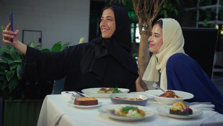 Expressions of happiness and pleasure at a gathering of friends, chatting and having a good time with friends, taking souvenir photos, a high-end coffee shop in the Emirates, developing social relations, two Arab Gulf Emirati women sitting at a table in a cafe, two Saudi friends exchanging talk.