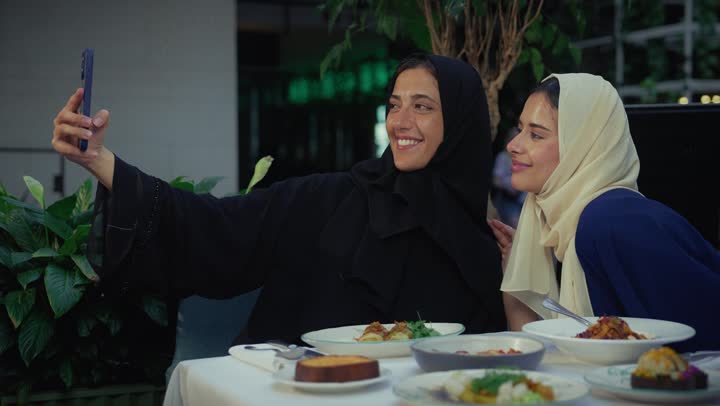 Chatting, having a good time with friends, taking souvenir photos, a classy coffee shop in the Emirates, developing social relationships, expressions of happiness and pleasure at a gathering of friends, two Arab Gulf Emirati women sitting at a table in a cafe, two Saudi friends exchanging talk.