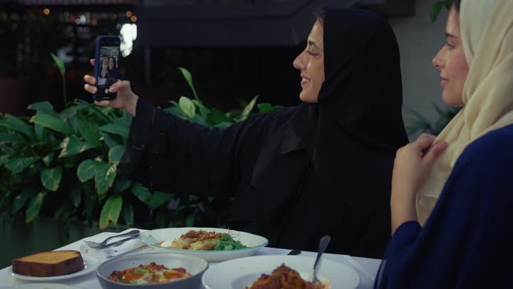التقاط السيلفي عبر الهاتف المحمول ، قضاء وقت ممتع مع الأصدقاء، تطوير العلاقات الإجتماعية، مشاركه الطعام مع الاصدقاء، توثيق الذكريات عبر الجوال ، صديقتان سعوديتين تتبادلان الحديث، كوفي شوب راقي بالامارات، امرأتان عربيتين خليجيتين اماراتيتين تجلسان على طاولة في المقهى ، تعابير السعادة والسرور بلمة الرفاق .