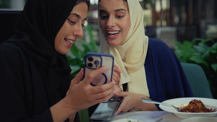 Documenting memories via mobile, two Saudi friends exchanging conversation, a high-end coffee shop in the Emirates, sharing food with friends, two Emirati Gulf Arab women sitting at a table in a cafe, developing social relationships, chatting and having a good time with friends, expressions of happiness and pleasure at a gathering of friends.
