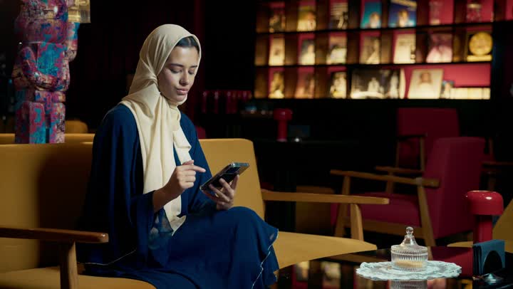 قضاء أوقات ممتعة ومسلية خارج المنزل، استعمال البرامج والتطبيقات الالكترونية، احتساء المشروب المفضل، تصوير لامراة عربية خليجية اماراتية محجبة ترتدي العباءة تجلس على الأريكة في إحدى المقاهي الراقية تستمع بلحظات الهدوء والراحة ، سيده سعودية تقوم بتصفح الهاتف المحمول