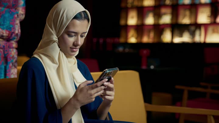 استعمال البرامج والتطبيقات الالكترونية، قضاء أوقات ممتعة ومسلية خارج المنزل، احتساء المشروب المفضل، تصوير لامراة عربية خليجية اماراتية محجبة ترتدي العباءة تجلس على الأريكة في إحدى المقاهي الراقية تستمع بلحظات الهدوء والراحة ، سيده سعودية تقوم بتصفح الهاتف المحمول