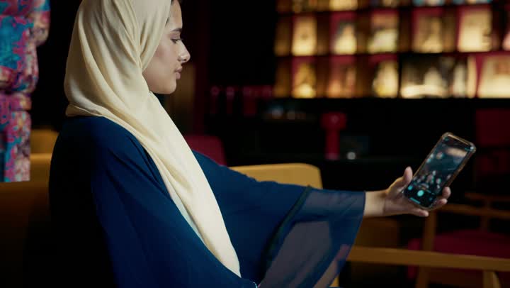 Online shopping, checking on family and friends, browsing social media, a smiling Emirati Gulf Arab woman wearing a black abaya sitting on a couch in an upscale cafe, a young Saudi woman browsing the internet on a tablet