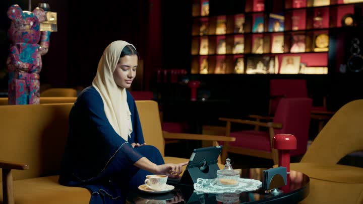 تصفح مواقع التواصل الاجتماعي، التسوق الالكتروني عبر الأونلاين، امراة عربيه خليجيه اماراتية مبتسمه ترتدي العباءه السوداء تجلس على الأريكة في إحدى المقاهي الرقية ، شابه سعودية تقوم تصفح الانترنت عبر جهاز التابلت