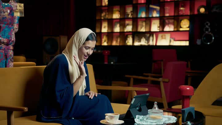 امراة عربيه خليجيه اماراتية مبتسمه ترتدي العباءه السوداء تجلس على الأريكة في إحدى المقاهي الرقية ، القاء التحيه والسلام ، التسوق الالكتروني عبر الأونلاين، الاطمئنان على الاهل والاصدقاء ، تصفح مواقع التواصل الاجتماعي،  شابه سعودية تقوم تصفح الانترنت عبر جهاز التابلت