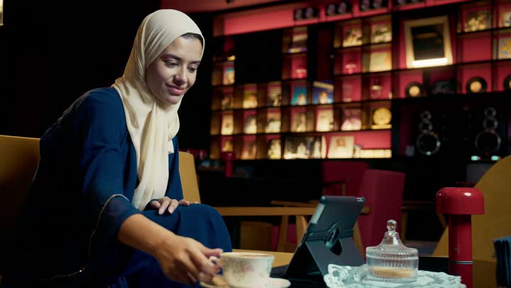 Having a good time, enjoying hot drinks, a smiling Emirati Gulf Arab woman wearing a black abaya sitting on a sofa in an upscale cafe, greeting people, online shopping, browsing social media, a young Saudi woman browsing the internet on a tablet