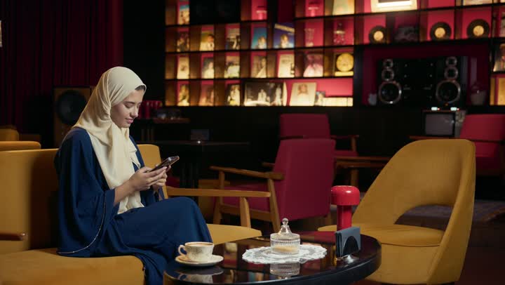 قضاء أوقات ممتعة ومسلية خارج المنزل،استعمال البرامج والتطبيقات الالكترونية، احتساء المشروب المفضل، تصوير لامراة عربية خليجية اماراتية محجبة ترتدي العباءة تجلس على الأريكة في إحدى المقاهي الراقية تستمع بلحظات الهدوء والراحة ، سيده سعودية تقوم بتصفح الهاتف المحمول