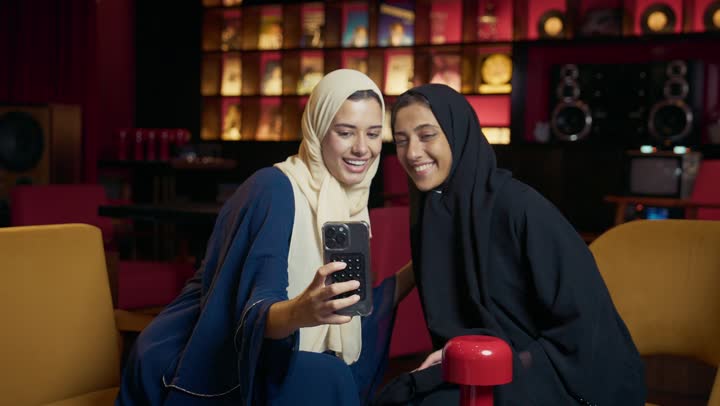 A classy coffee shop in the Emirates, developing social relationships, chatting and having a good time with friends, taking souvenir photos, expressions of happiness and pleasure at the gathering of friends, two Arab Gulf Emirati women sitting at a table in the cafe, two Saudi friends exchanging conversation.