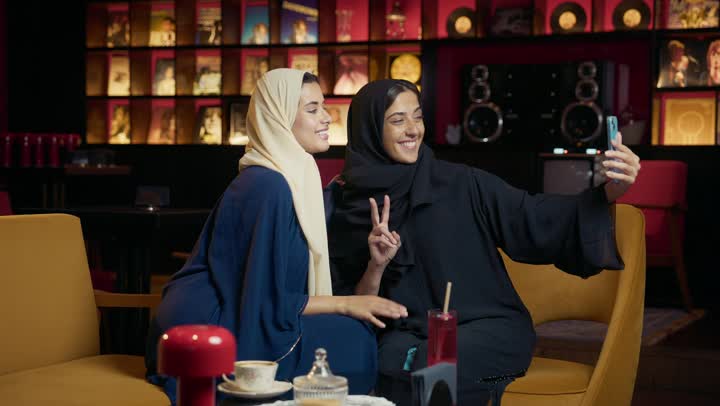 Chatting, having a good time with friends, taking souvenir photos, a classy coffee shop in the Emirates, developing social relationships, expressions of happiness and pleasure at a gathering of friends, two Arab Gulf Emirati women sitting at a table in a cafe, two Saudi friends exchanging talk.