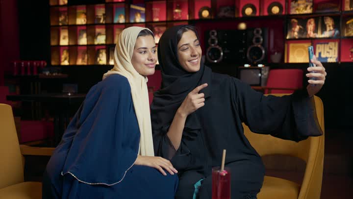 التقاط الصور التذكارية ، كوفي شوب راقي بالامارات، تطوير العلاقات الإجتماعية، التسامر والدردشة وقضاء وقت ممتع مع الأصدقاء، تعابير السعادة والسرور بلمة الرفاق، امرأتان عربيتين خليجيتين اماراتيتين تجلسان على طاولة في المقهى، صديقتان سعوديتين تتبادلان الحديث .