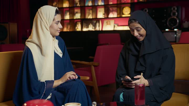 قضاء وقت ممتع مع الأصدقاء ، صديقتان سعوديتين تتبادلان الحديث ، استخدام التكنولوجيا والتقنيات الحديثه ، كوفي شوب راقي بالامارات، تطوير العلاقات الإجتماعية، تعابير السعادة والسرور بلمة الرفاق، امرأتان عربيتين خليجيتين اماراتيتين تجلسان على طاولة في المقهى.