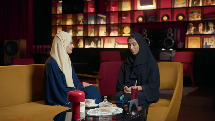 Gestures of happiness and enthusiasm, two Saudi friends exchanging talk, using technology and modern techniques, a high-end coffee shop in the Emirates, developing social relations, chatting and spending quality time with friends, expressions of happiness and pleasure at a gathering of friends, two Arab Gulf Emirati women sitting at a table in a cafe.