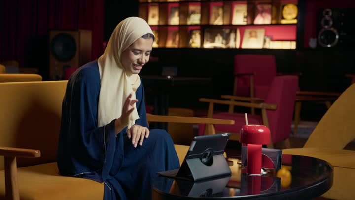 دمج التكنولوجيا المتطورة في الحياة اليومية، التواصل مع الأهل والأصدقاء، احتساء المشروب المفضل، امراة عربية خليجية اماراتية محجبة ترتدي العباءة تجلس على الأريكة في إحدى المقاهي الراقية تستمع بلحظات الهدوء والراحة ، سيده سعودية تقوم باجراء مكالمة فيديو عبر التابلت