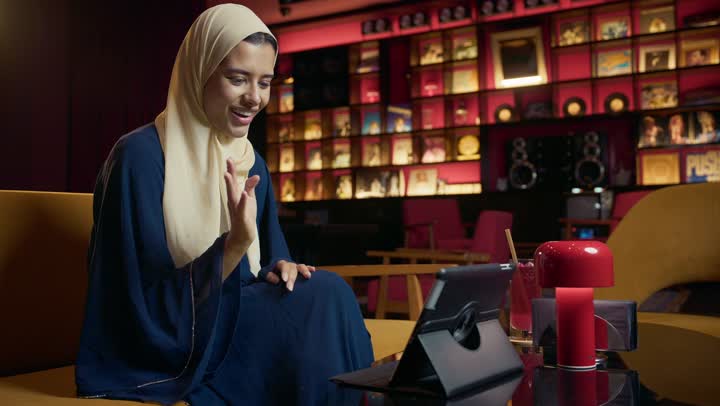 التواصل مع الأهل والأصدقاء، دمج التكنولوجيا المتطورة في الحياة اليومية، احتساء المشروب المفضل، امراة عربية خليجية اماراتية محجبة ترتدي العباءة تجلس على الأريكة في إحدى المقاهي الراقية تستمع بلحظات الهدوء والراحة ، سيده سعودية تقوم باجراء مكالمة فيديو عبر التابلت