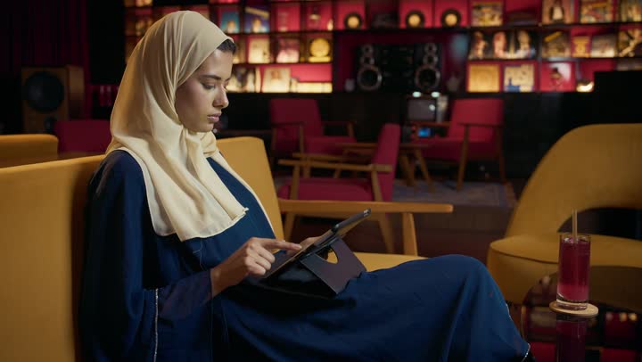 سيده سعودية تقوم باجراء مكالمة فيديو عبر التابلت ، التواصل مع الأهل والأصدقاء، دمج التكنولوجيا المتطورة في الحياة اليومية،  امراة عربية خليجية اماراتية محجبة ترتدي العباءة تجلس على الأريكة في إحدى المقاهي الراقية تستمع بلحظات الهدوء والراحة