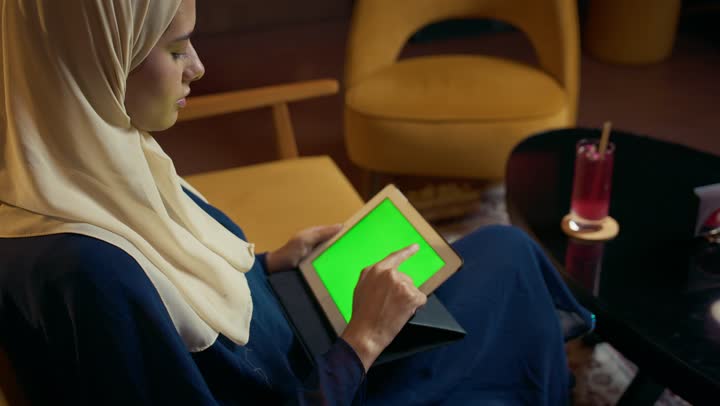 عرض شاشه خضراء ، امراة عربية خليجية اماراتية محجبة ترتدي العباءة تجلس على الأريكة في إحدى المقاهي الراقية تستمع بلحظات الهدوء والراحة ، سيده سعودية تقوم باجراء مكالمة فيديو عبر التابلت ، التواصل مع الأهل والأصدقاء، دمج التكنولوجيا المتطورة في الحياة اليومية