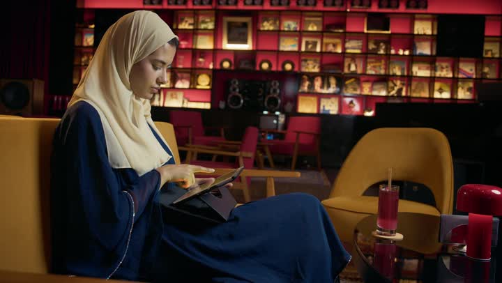 التواصل مع الأهل والأصدقاء، عرض شاشه خضراء ، امراة عربية خليجية اماراتية محجبة ترتدي العباءة تجلس على الأريكة في إحدى المقاهي الراقية تستمع بلحظات الهدوء والراحة ، سيده سعودية تقوم باجراء مكالمة فيديو عبر التابلت ، دمج التكنولوجيا المتطورة في الحياة اليومية