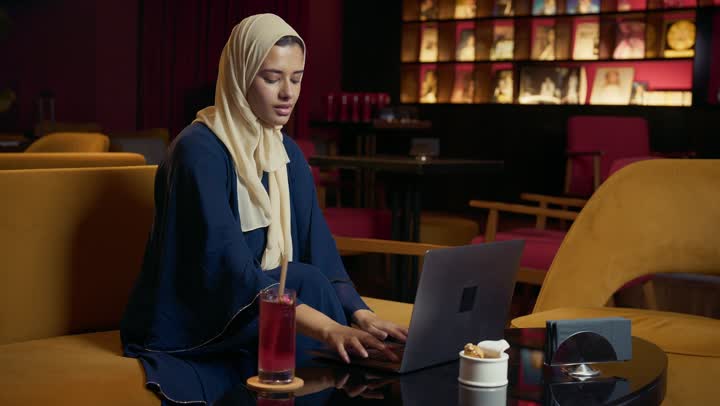 تطوير الاعمال والاستراتيجيات, النجاح وتحقيق الاهداف، التواصل مع الأهل والأصدقاء، استخدم جهاز الحاسوب المحمول ، امراة عربية خليجية اماراتية محجبة ترتدي العباءة تجلس على الأريكة في إحدى المقاهي الراقية تستمع بلحظات الهدوء والراحة ، سيده سعودية تقوم بانجاز المشاريع والمهام في الكافيه.