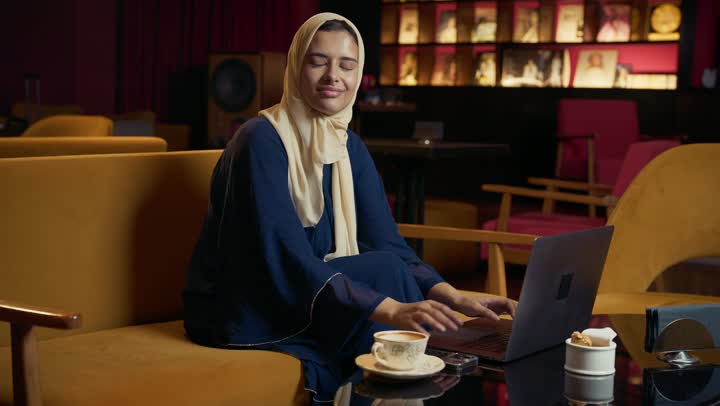استخدم جهاز الحاسوب المحمول ، النجاح وتحقيق الاهداف، تطوير الاعمال والاستراتيجيات, التواصل مع الأهل والأصدقاء، امراة عربية خليجية اماراتية محجبة ترتدي العباءة تجلس على الأريكة في إحدى المقاهي الراقية تستمع بلحظات الهدوء والراحة ، سيده سعودية تقوم بانجاز المشاريع والمهام في الكافيه.