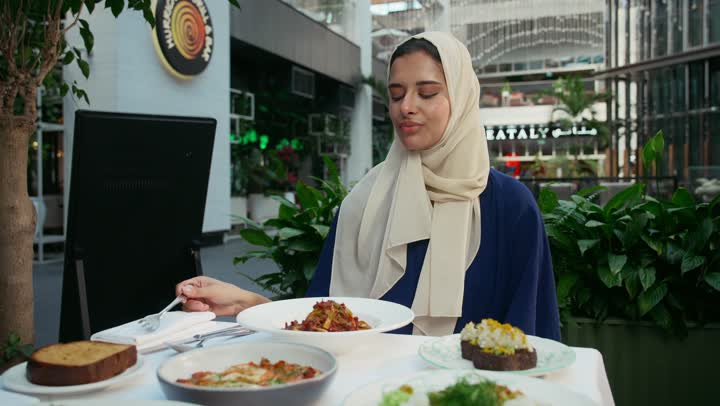 مطاعم و كافيهات في الإمارات ، تناول الغداء في المطعم, الاستمتاع بالأنشطة الترفيهية, امرأة عربية خليجية إماراتية محجبة ترتدي الزي الكاجوال تجلس على المائدة و تنظر إلى الكاميرا بإيماءات السعادة و السرور, قضاء الأوقات الممتعة في الخارج .
