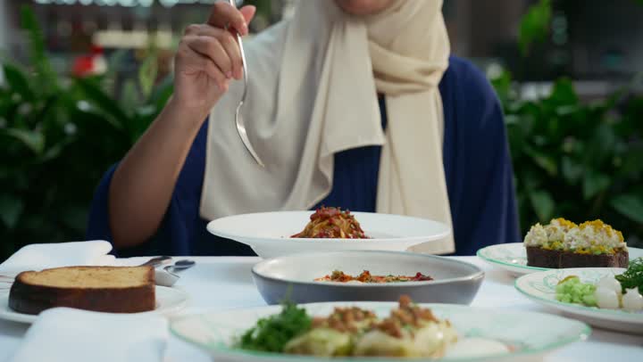 Having lunch at a restaurant, restaurants and cafes in the Emirates, enjoying recreational activities, a veiled Emirati Gulf Arab woman wearing casual clothes sitting at a table and looking at the camera with gestures of happiness and pleasure, spending a pleasant time outside.