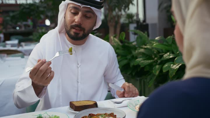 Affection and intimacy between spouses, a table full of delicious food, a close-up of an Emirati Gulf Arab couple sitting at the table, restaurants and cafes in the Emirates, gestures of happiness and pleasure, enjoying recreational activities, spending fun times outside, having lunch at a restaurant
